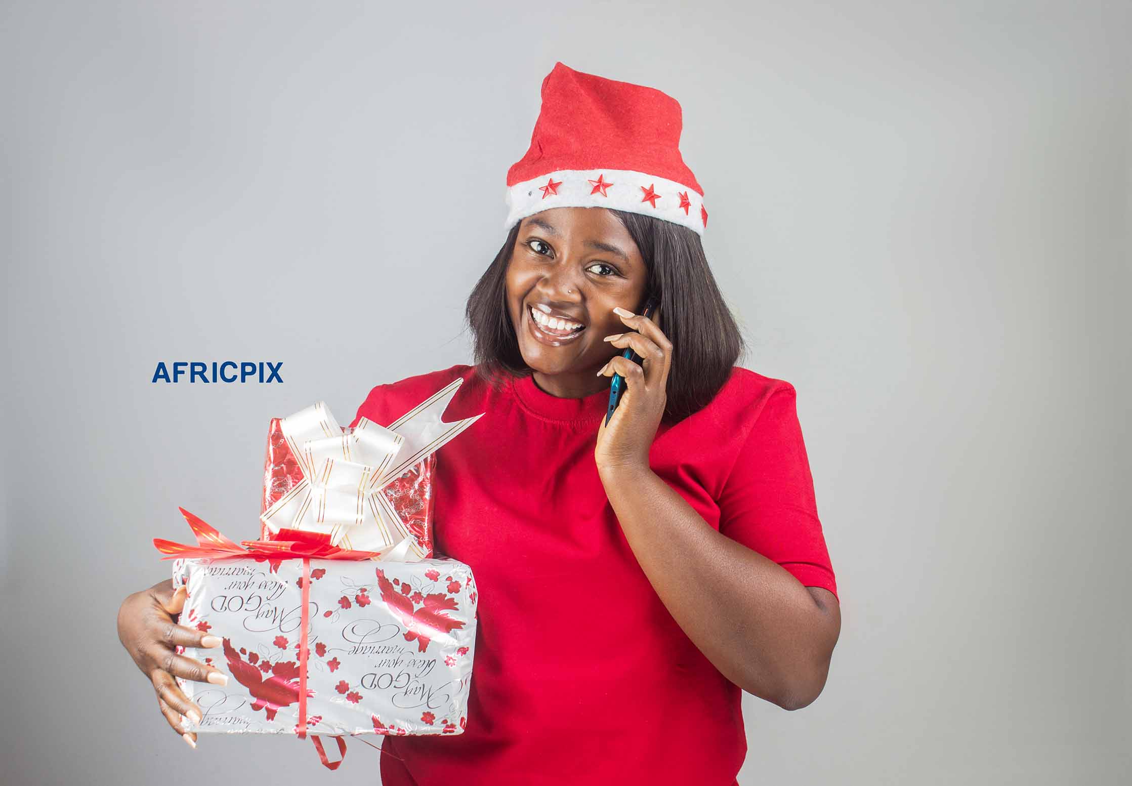 A happy African Nigerian woman wearing a Christmas cap, smiling while making a call and holding a gift box.